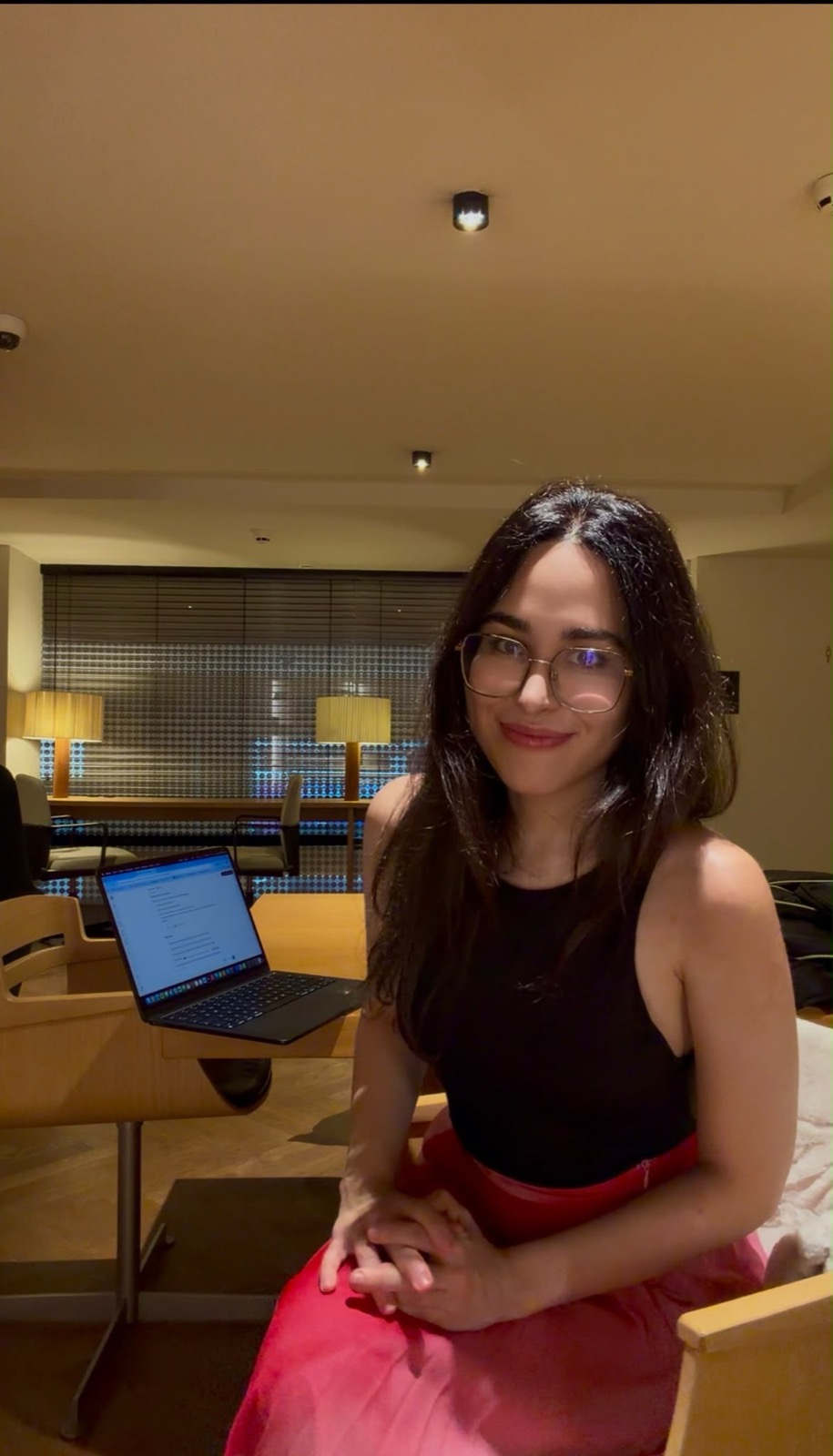 Clara Agnes wearing glasses and a black top, looking directly at the camera with a warm, open smile in a softly lit room