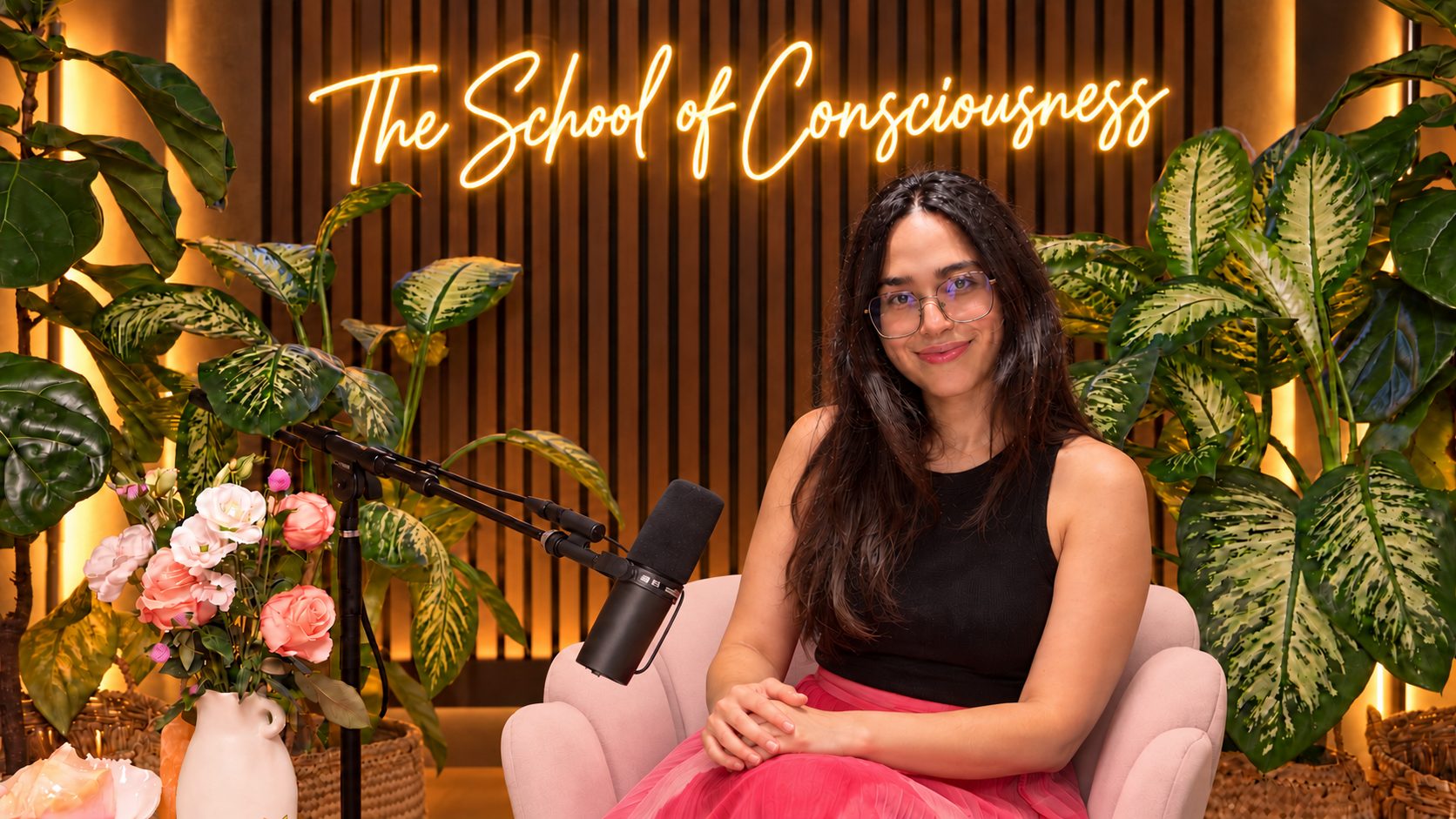 Clara Agnes seated at the podcast microphone with her hands clasped and an open, warm smile, the neon sign reading The School of Consciousness glowing on the wood-panelled wall behind her
