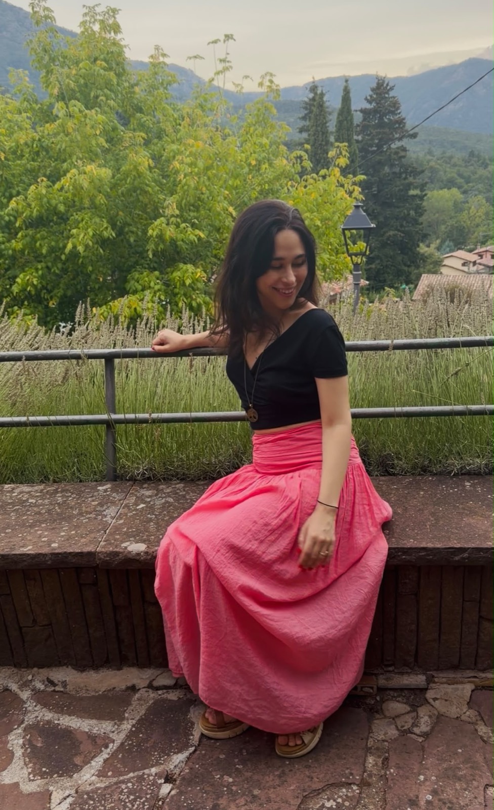 Clara Agnes in a bright pink skirt and black top, sitting outdoors by a lavender field with green hills behind her, looking down with a soft smile